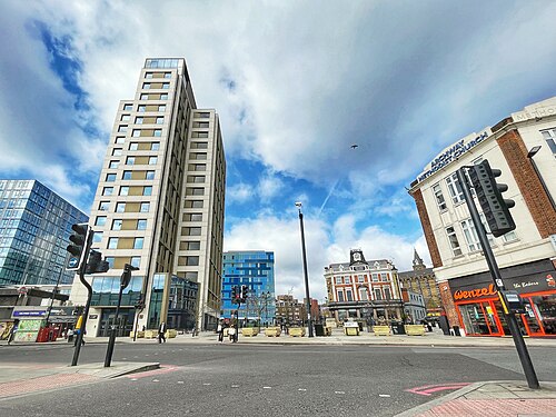 Wide city street with a tall modern apartment building on the left, smaller shops on the right, and a bright blue sky with clouds above.
